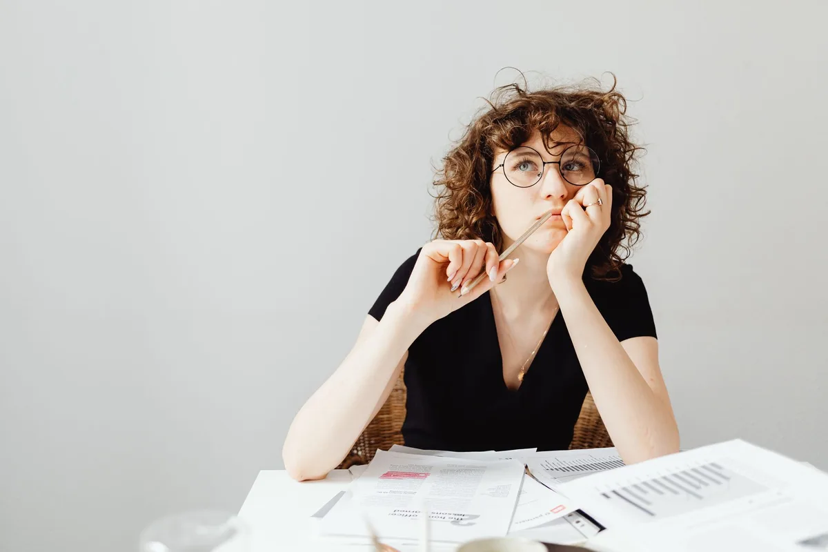 A woman with glasses thinking carefully at a desk covered in documents — the face of analysis paralysis in startup planning