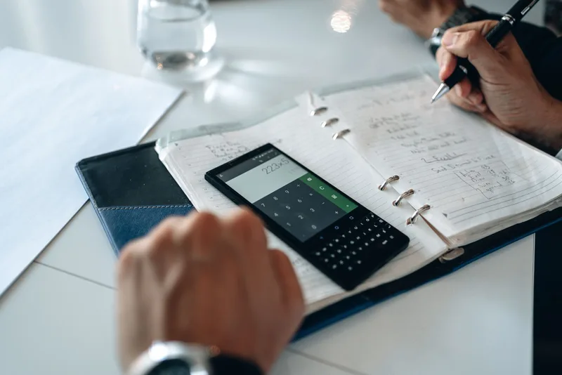 Close-up of hands writing in a notebook next to a smartphone calculator on a desk