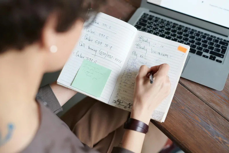 A person at a desk reading Reddit threads on a laptop, taking notes in a paper notebook